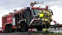 Eine junge Frau in hellbrauner Feuerwehruniform steht vor einem Löschfahrzeug.