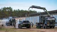 Soldaten in ABC-Schutzausrüstung reinigen mit Sprühpistolen Militärfahrzeuge neben einer Lagerhalle