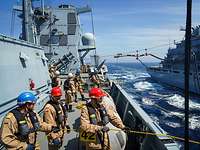 Mehrere Soldaten stehen auf dem Seitendeck eines Schiffes. Im Hintergrund wird eine Schlauchverbindung errichtet