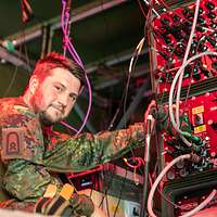 Ein Soldat posiert vor der Rückseite eines IT-Geräts mit zahlreichen angesteckten Kabeln fürs Foto 