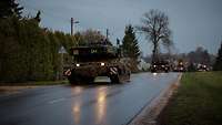 Mehrere Kampfpanzer sowie Schützenpanzer fahren hintereinander auf einer öffentlichen Straße