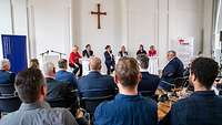 5 Frauen und ein Mann sitzen auf einem Podium unter einem Kreuz und diskutieren vor anderen Menschen