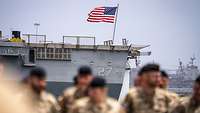 Soldaten stehen auf einem Schiff. Im Hintergrund weht die US-amerikanische Flagge.