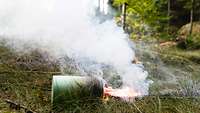 A round green can is lying on the ground in a forest. A flame and smoke are flowing out sideways.