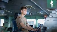 Ein Soldat sitzt an einem Steuerrad auf der Brücke eines Kriegsschiffes.