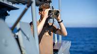 Ein Soldat schaut von Deck eines Schiffes mit einem Fernglas aufs Meer hinaus.