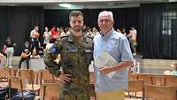 Ein Soldat und ein Zivilist posieren in einem Saal für ein Foto. Im Hintergrund sind mehrere Zivilisten zu sehen.