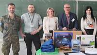 Zwei Soldaten und vier Zivilisten stehen aufgereiht vor einer Tafel. Davor ein Tisch mit Spenden.