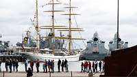 Besucher gehen an einem weißen Segelschiff vorbei
