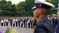 Soldaten und Zivilisten stehen im Halbkreis. Einige von ihnen halten Blumenkränze