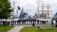 Eine lange Menschenschlange bildete sich in einem Hafen. Im Hintergrund ist ein Segelschiff.