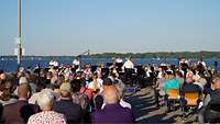 Ein Musikchor spielt vor einem weitläufigen Gewässer
