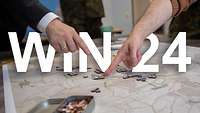 Eine Wargame-Karte liegt auf dem Tisch. Zwei Finger zeigen auf Spielsteine. Darauf steht WIN 24.