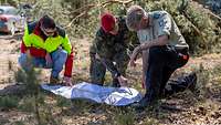 Ein Soldat kniet mit zwei weiteren Personen auf einem Waldboden vor einer Landkarte