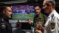 Drei Personen in Uniform von der Polizei, Flugsicherung und Bundeswehr stehen vor einem Bildschirm auf dem Fußball läuft