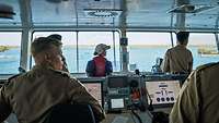 Marinesoldaten in Arbeitsuniform und eine Zivilistin in Arbeitsjacke auf einer Schiffsbrücke