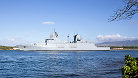 Ein graues Kriegsschiff in See vor einer hügeligen, grünen Küste.