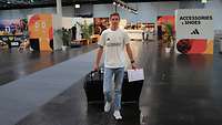 Ein Mann im Olympia-Trikot läuft mit zwei schwarzen Rolltaschen und einem Formular durch eine Halle.