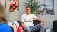 Ein Mann sitzt auf einem Sofa neben dem Olympia-Maskottchen. Dahinter ein Bild vom Eiffelturm.
