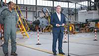 Ein Zivilist steht in einer Halle an einem Mikrofon und spricht. Im Hintergrund stehen Kampfjets
