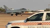In the background there is a fighter jet while in the foreground there is an air traffic control car.