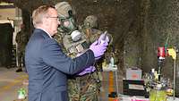 Ein Mann im Anzug und ein Soldat mit ABC-Schutzmaske halten ein Laborgerät in den Händen und blicken darauf