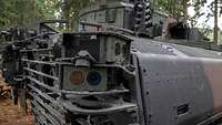 Großaufnahme von Optiken hinten rechts an einem Panzer, der in einem Waldstück steht