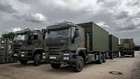 Mehrere schwere Lkw mit großen grünen Containern stehen auf einer Betonfläche.