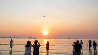 Während die Sonne über dem Meer untergeht fliegt ein Jet übers Wasser 
