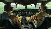 Zwei Piloten sitzen im Cockpit. Aus dem Fenster sieht man eine grüne Landschaft. 