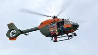 Ein Hubschrauber im Schwebeflug. Drei Personen stehen an der geöffneten Helikoptertür.