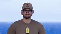 Ein Mann mit Brille schaut in die Kamera, im Hintergrund verschwimmt das Meer.