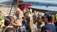 A group of soldiers is standing around a soldier, with a combat aircraft in the background.