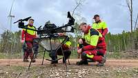 Vier Männer knien vor einer Drohne die einen Trichter unter sich trägt auf einem Waldweg