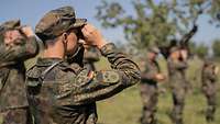 Mehrere Soldaten stehen im Halbkreis und schauen ins Gelände vor ihnen durch Ferngläser.