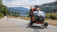 Ein Rettungshubschrauber vom Typ SAR H145M steht auf einem Schotterplatz zwischen Wasser und Gebirge