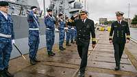 Zwei Männer in dunkler Marineuniform schreiten eine Formation mit präsentierenden Gewehren ab.
