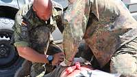 Zwei Soldaten behandeln einen Verletzten am Boden. Auf seinem Bauch sind viele Tücher mit Kunstblut angebracht. 