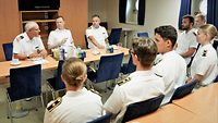Ein Marineoffizier sitzt hinter einem Tisch und spricht in eine Runde aus Marinesoldaten.