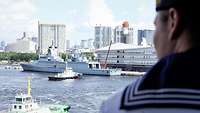 Ein graues Schiff liegt in einem Hafen. Im Hintergrund sind Hochhäuser zu sehen.