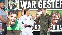 Eine Judoka spricht mit einem Soldaten, daneben ein Trainer. Im Hintergrund ein Bildschirm.
