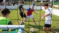 Blick in Kleinfußballfeld über Schulter des Torwarts. Davor drei junge Männer im Kampf um den Ball.