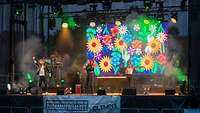 Blick auf eine Bühne mit DJ und Musikern. Im Hintergrund ein Blumenmotiv auf großem Bildschirm.