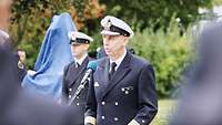 Ein Soldat in Marineuniform spricht in ein Mikrofon. Hinter ihm befinden sich weitere Soldaten und eine abgedecktes Objekt.