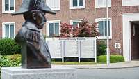 Im Vordergrund eine Büste eines Marineadmirals aus Bronze. Im Hintergrund ein Schild mit dem Namen der Liegenschaft.