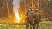Zwei Personen in Flecktarnuniform stehen vor einem Lagerfeuer im Wald