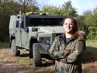 Eine Frau in Uniform und Brille steht vor einem olivgrünem gepanzerten Fahrzeug