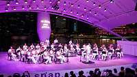 Ein Orchester bestehend aus vielen weiß uniformierten Personen spielt ein Open-Air-Konzert.