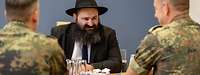 Ein Rabbiner mit schwarzem Hut unterhält sich an einem Tisch sitzend mit zwei Soldaten in Flecktarn