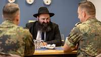 Ein Rabbiner mit schwarzem Hut unterhält sich an einem Tisch sitzend mit zwei Soldaten in Flecktarn